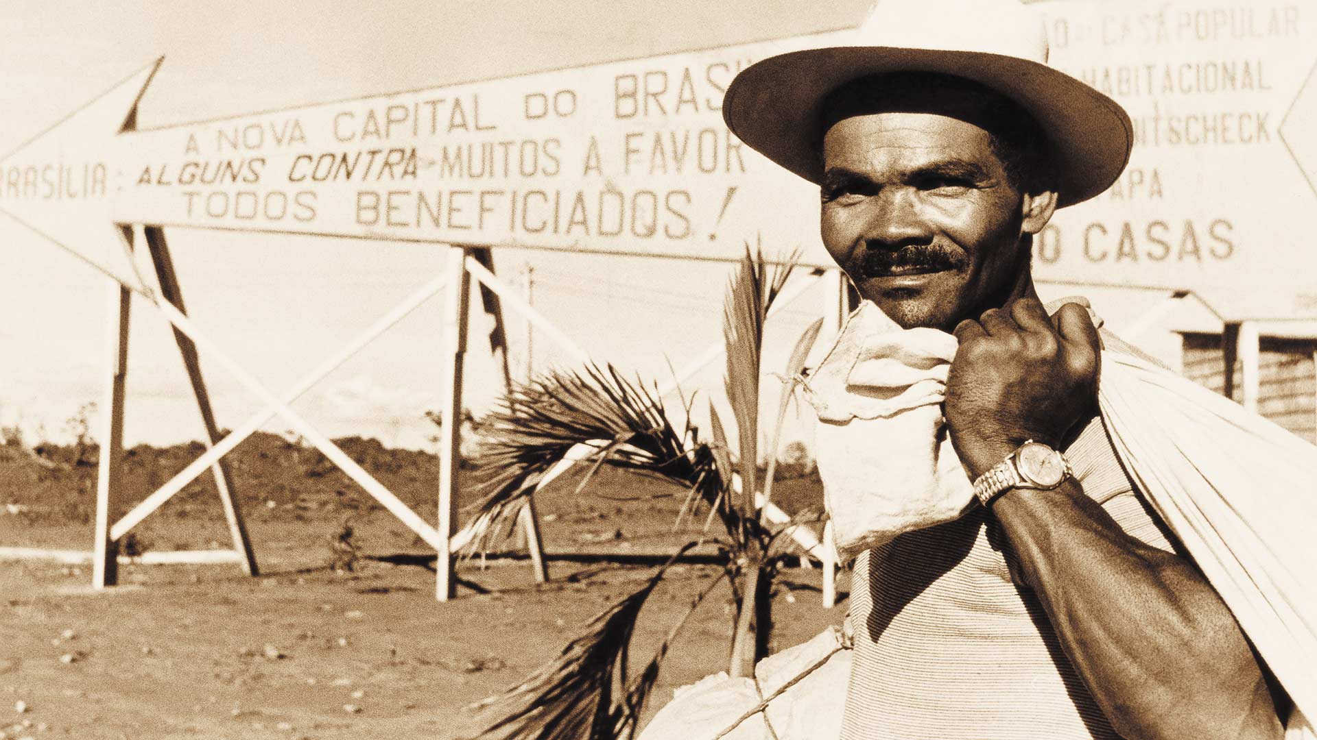 Fotografia envelhecida de Åke Borglund. À direita está um homem negro enquadrado do tórax para cima. Ele usa chapéu, tem rugas na testa, olhos pequenos, nariz largo, bigode e um leve sorriso. Ele usa camisa clara de manga curta com listras finas horizontais. Ele carrega sobre o ombro esquerdo um saco de pano e usa relógio. Ao fundo há uma placa em formato de seta, parcialmente encoberta pelo homem onde se vê escrito: “Brasília: A nova capital do Brasil. Alguns contra - muitos a favor. Todos beneficiados!”