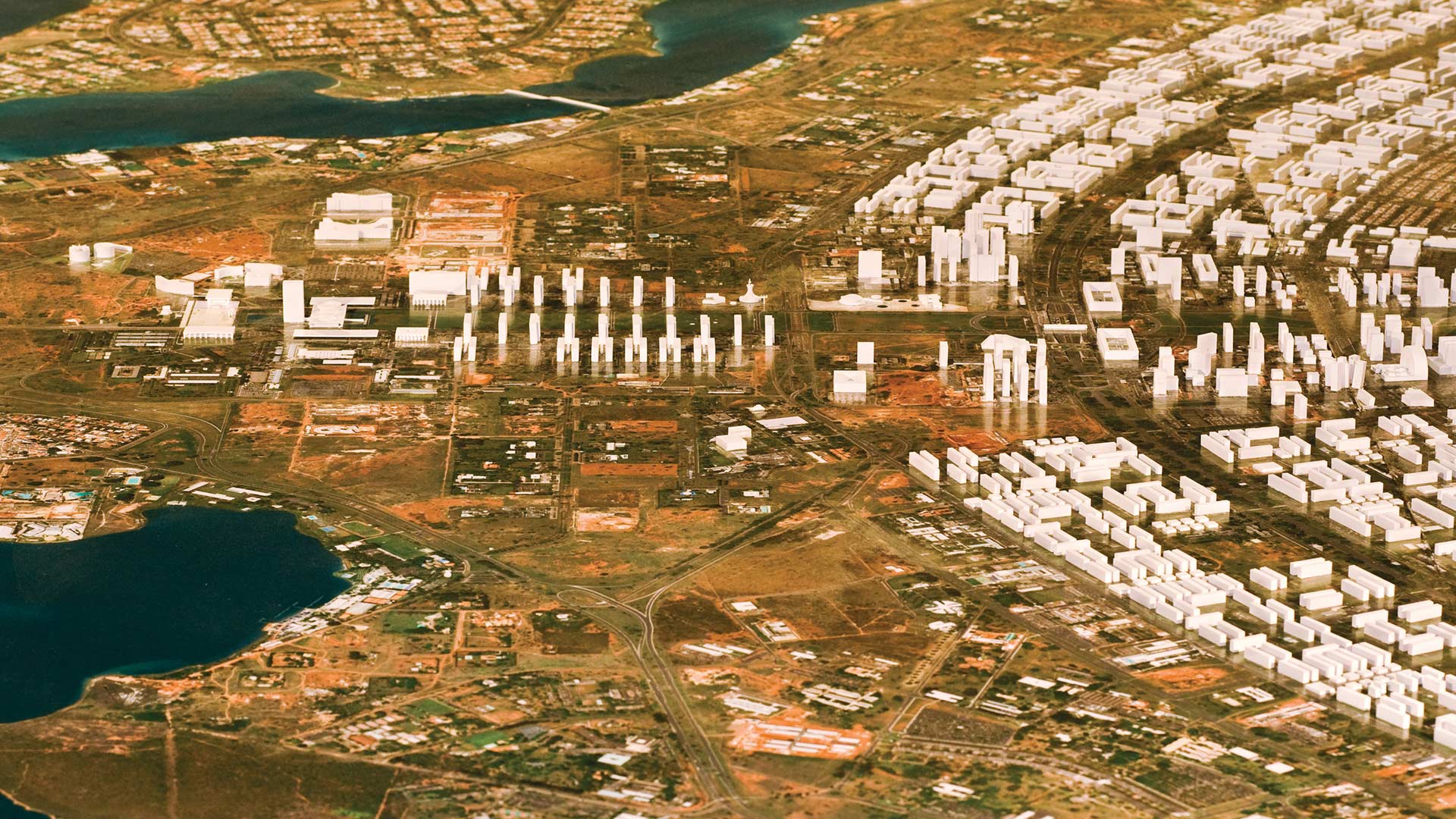 Fotografia colorida da maquete de Brasília, feita pelo arquiteto Antônio José de Oliveira. À esquerda está o Lago Paranoá pintado de azul, no centro toda Esplanada dos Ministérios com todas as edificações representadas por miniaturas pintadas de branco. Também é possível ver todas as ruas e vias de acesso. À direita estão as miniaturas dos prédios das Asa Sul e Norte.