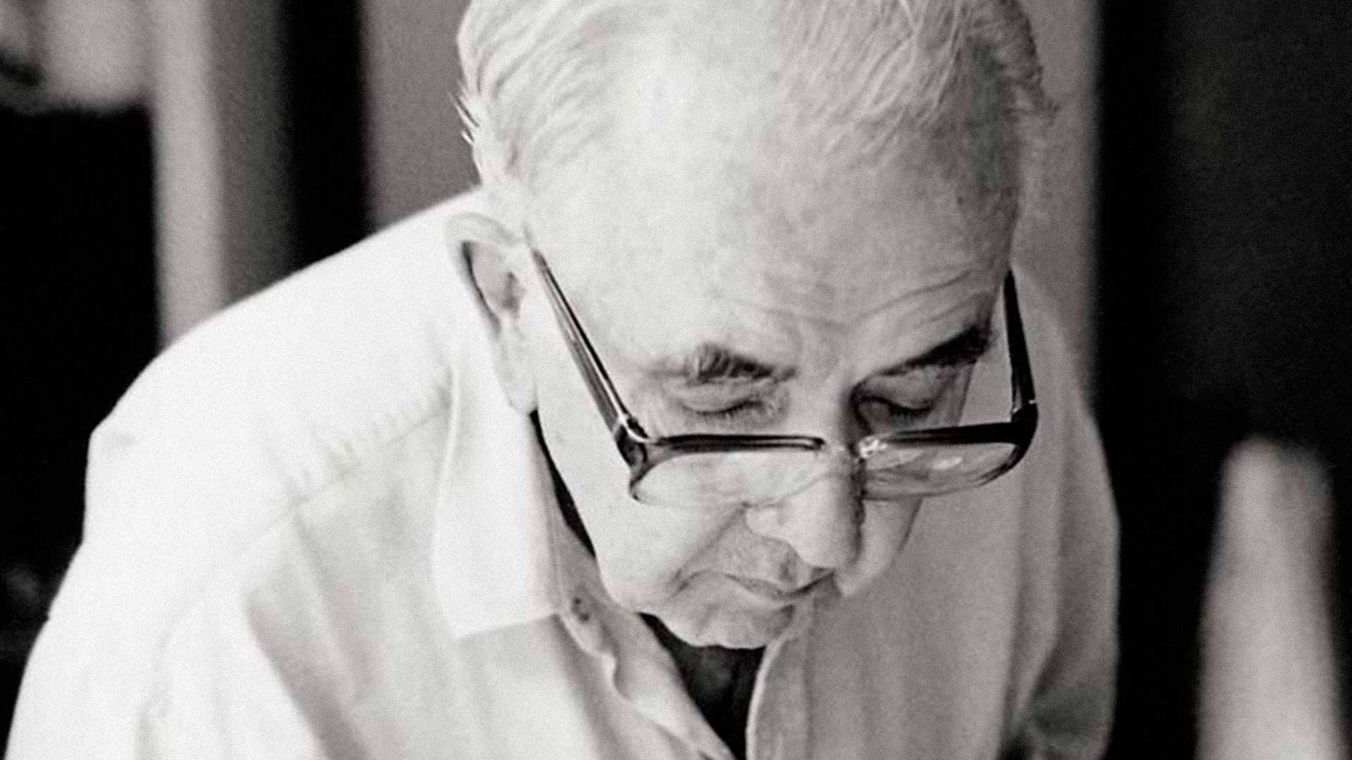 Fotografia em preto e branco de Athos Bulcão. Ele aparece sentado e curvado como se estivesse lendo algo. Ele tem cabelos brancos, sobrancelhas grossas e pretas, nariz fino e boca pequena, usa óculos com armação preta e lentes grossas.