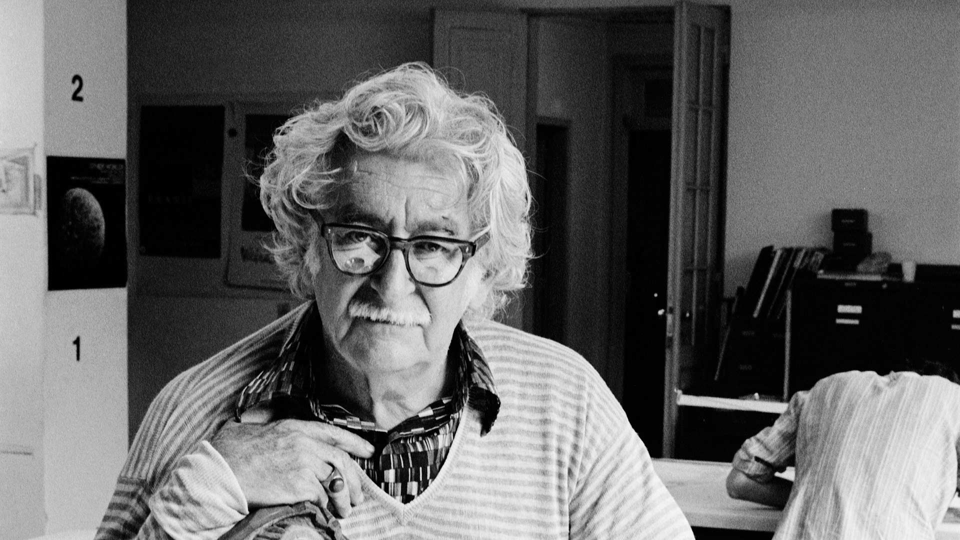 Fotografia em preto e branco de Roberto Burle Marx. Ele está enquadrado do tórax para cima, sentado. Ele tem pele clara, seus cabelos brancos estão bagunçados, tem sobrancelhas grossas, usa óculos com armação escura e na lente há o reflexo da mesa à sua frente. Tem nariz grande, usa bigode ralo e tem lábios finos. Ele veste camisa estampada com colarinho aberto. Sobre essa camisa ele veste uma blusa de gola em “v” com listras na horizontal claras e finas.  Ao fundo há uma porta aberta, uma pessoa de costas em frente a uma mesa com vários livros e pequenas gavetas. Nas paredes há posters.