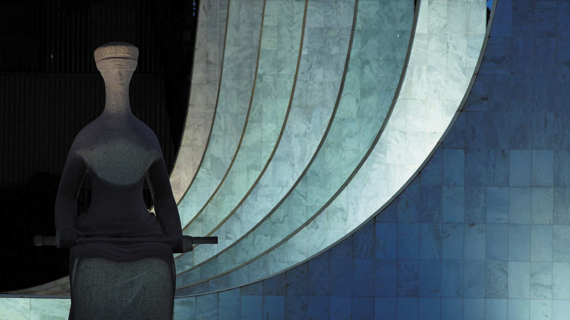 Fotografia de Fabio Colombini da Estátua do Palácio da Justiça em preto e branco. Em destaque à esquerda da tela está a Estátua da Justiça, que é representada por uma mulher, sentada e vendada, sobre seu colo ela segura uma espada. Ao fundo do lado direito da tela 6 pilares curvos que fazem o desenho arquitetônico do prédio do Palácio do Supremo Tribunal Federal.