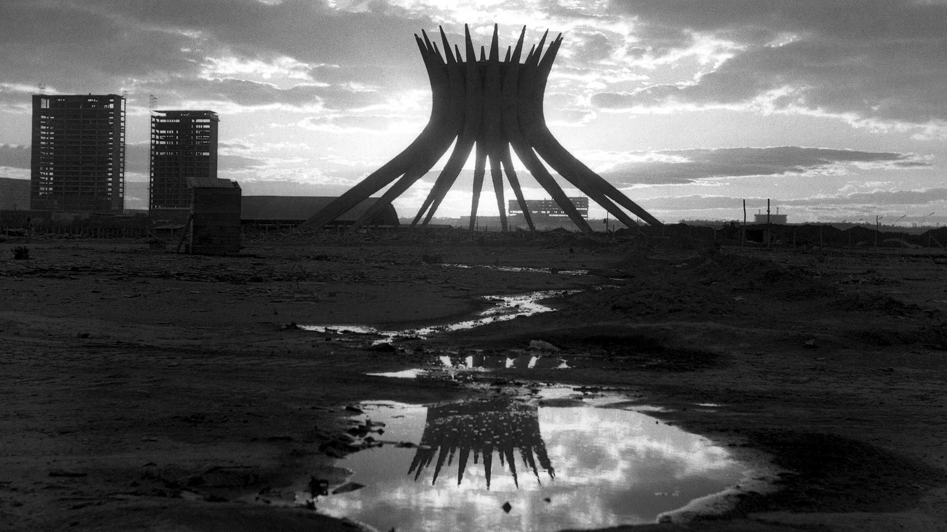 Fotografia em preto e branco de Gabriel Gondim. Em destaque ao centro uma poça de água reflete a estrutura da Catedral que está ao fundo realçada pelo reflexo da luz do sol e do céu coberto de nuvens. Mais distantes estão os canteiros de obras e à esquerda os esqueletos dos prédios dos futuros Ministérios.