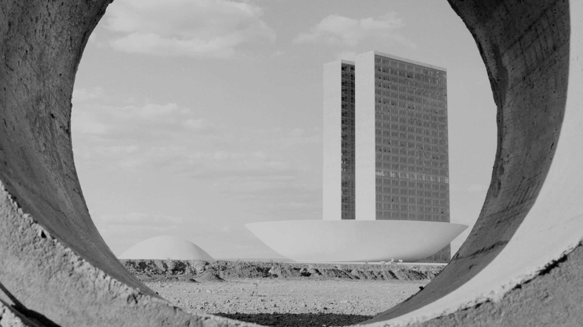 Fotografia em preto e branco de Jesco Puttkamer tirada por trás do arco de uma manilha de concreto. Mostra o Congresso Nacional em construção. Ao fundo é possível ver as cúpulas semi-esféricas do Senado Federal e da Câmara dos Deputados além das duas torres centrais.