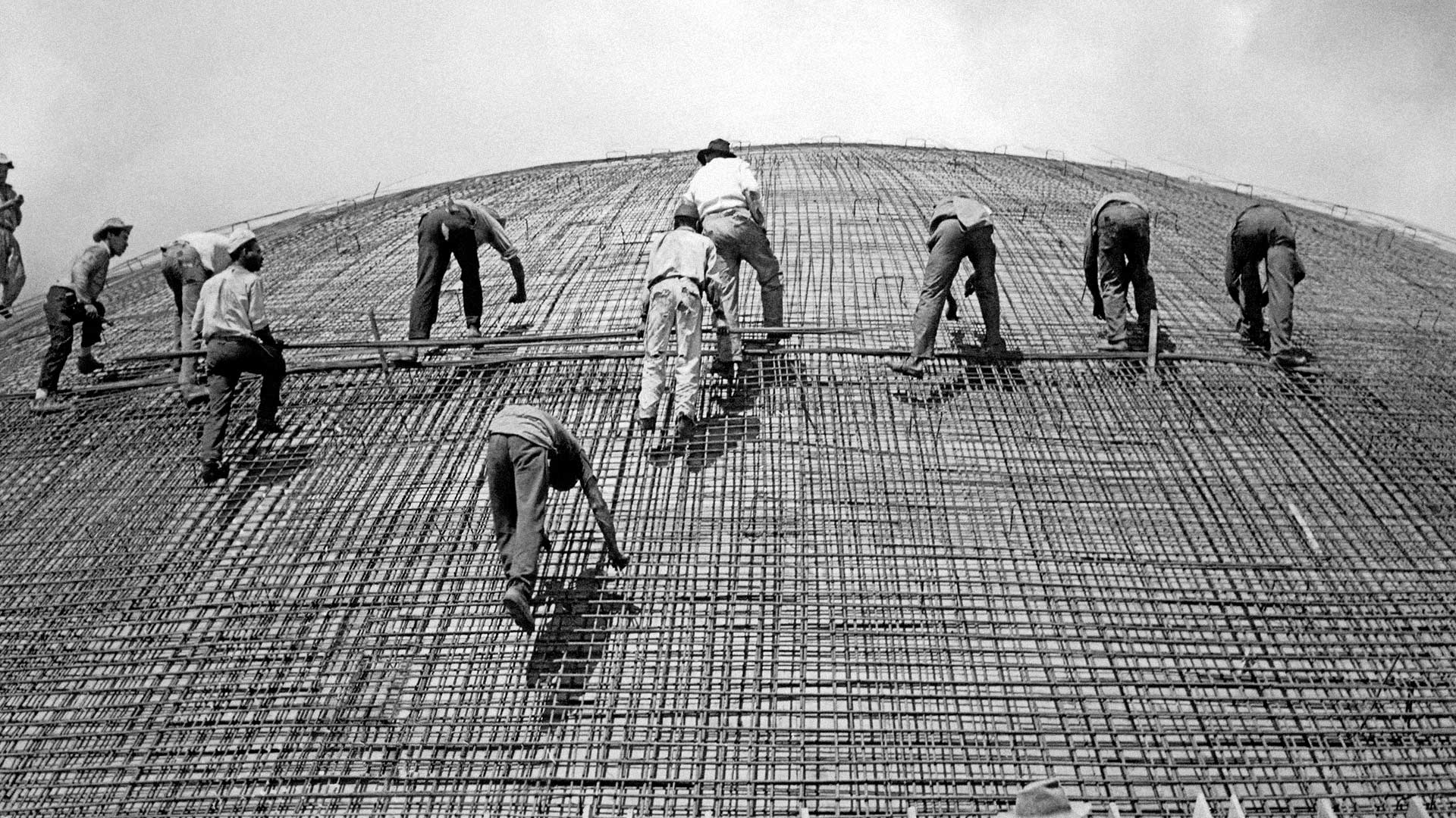 Fotografia em preto e branco de Marcel Gautherot mostra 11 trabalhadores que estão sobre um aramado metálico sobreposto a estrutura esférica do Senado Federal em construção. Eles estão de costas, usam chapéus, camisas de mangas longas e calças compridas.