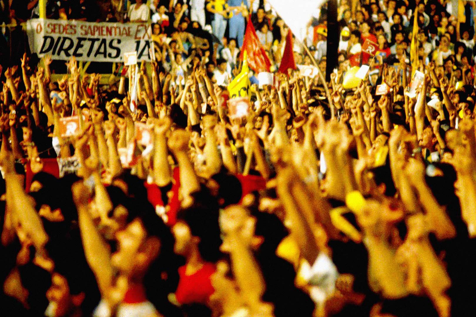 Fotografia de Ulysses Guimarães que tem pele clara, é calvo e os poucos cabelos que aparecem na lateral de sua cabeça são brancos. Veste terno azul escuro e uma camisa de manga comprida azul clara.  Ao fundo aparece céu cinza e a imagem do Congresso Nacional meio desfocada, em primeiro plano ele caminha com as mãos para trás e a cabeça ligeiramente baixa pelo gramado verde da direita para a esquerda, aparecendo de lado na tela.