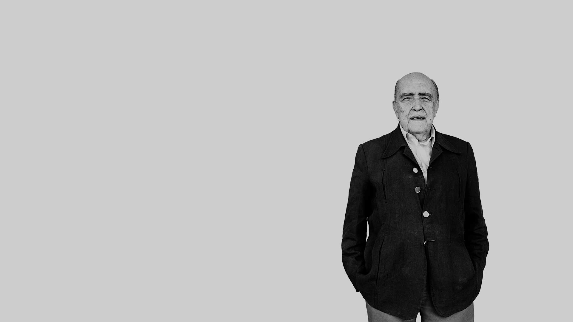 Fotografia em preto e branco de Oscar Niemeyer. Ele está do lado direito da tela de pé com as mãos nos bolsos da calça. Ele tem pele clara, é calvo, tem poucos cabelos escuros na lateral da cabeça, seus olhos são pequenos e sua boca está entreaberta. Em seu rosto há várias manchas escuras. Está vestindo casaco escuro de 4 botões grandes, mas só um está fechado. Por dentro há uma camisa clara com o colarinho aberto.