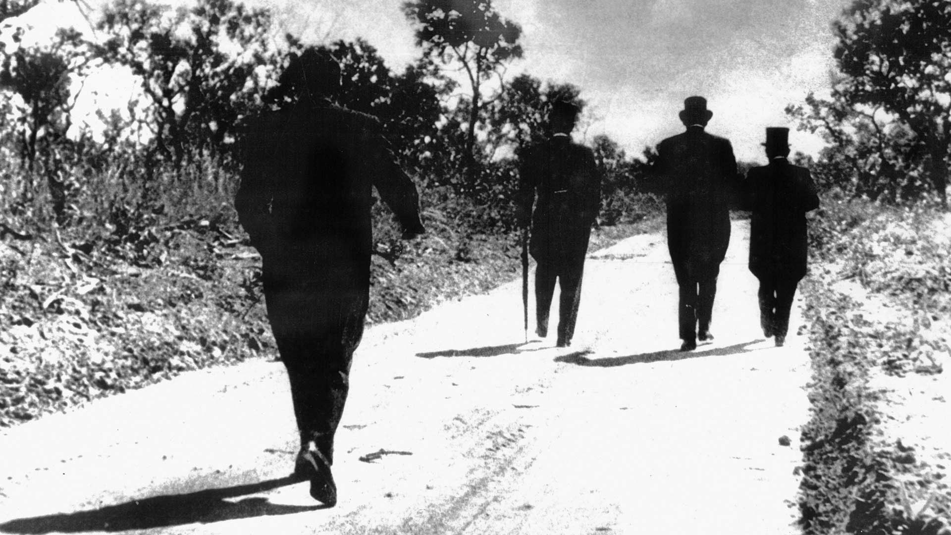 Fotografia em preto e branco de Raymond Frajmund onde se vê 4 homens caminhando por uma estrada de terra batida e nas laterais uma vegetação rasteira de cerrado. Eles aparecem de costas e todos vestem ternos sociais completos pretos e cartolas e um deles usa bengala de apoio.