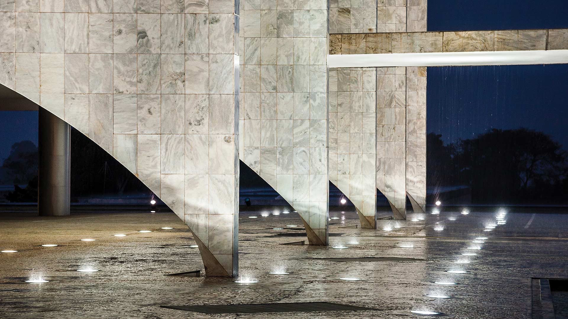 Fotografia de Rui Faquini de parte inferior do Palácio do Planalto. Foto noturna mostra 5 pilares que tem uma base estreita e a parte superior é mais larga na forma triangular invertida em forma de “V” (tendo formas retílíneas e geometricas), revestidas de mármore branco. O piso também em mármore com a evidência de 5 caminhos de luzes brancas iluminando o chão. Ao fundo céu azul escuro com sombras de árvores 