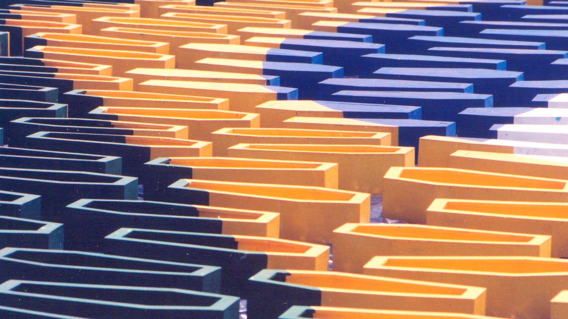 Fotografia parcial da instalação de Siron Franco nomeada “Caixões” apresenta caixões abertos, próximos à extremidade e fechados, mais ao centro, pintados de amarelo, azul e branco, um ao lado do outro, formando a bandeira do Brasil.